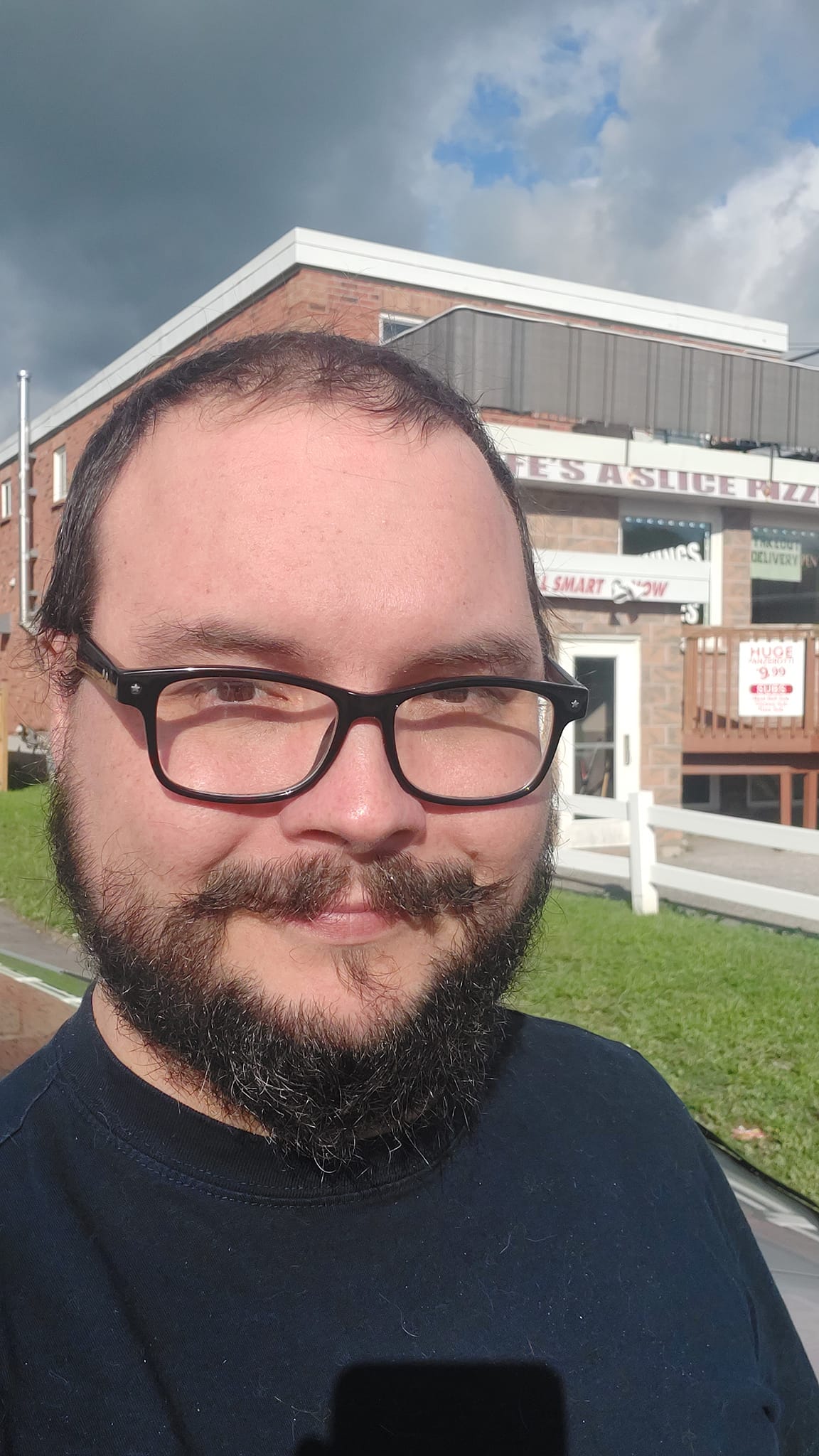 A man short hair and bushy facial hair stands in front of a small town pizzaria called Life's a Slice Pizza